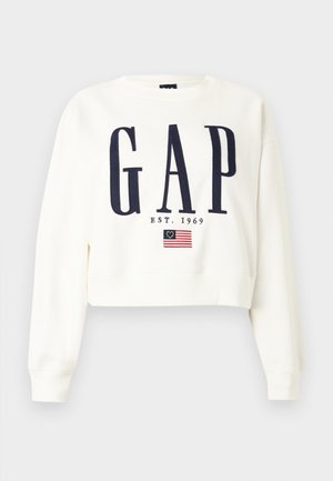 Bijela skraćena majica dugih rukava s velikim tamnoplavo natpisom "GAP", ispod njega "EST. 1969" i malom grafikom američke zastave Betsy Ross.