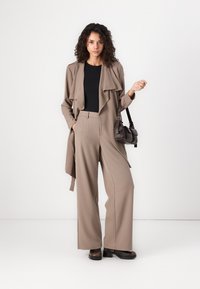 Beige, lettr trenchcoat med draperet krave over en sort top, parret med højtaljede, brede bukser og brune klodsede hæle.