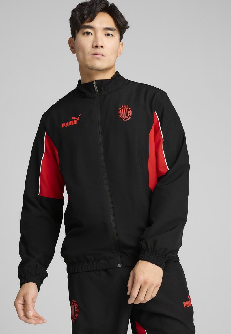 Črna jakna na zadrgo z rdečimi poudarki, ki ima elastične manšete in pas. Puma logotip in grb na prsih. Gladka tekstura materiala.