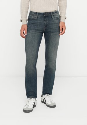 Jeans da uomo in denim blu scuro con vestibilità slim, leggere sfumature, design classico a cinque tasche e vita media, abbinati a sneakers bianche.