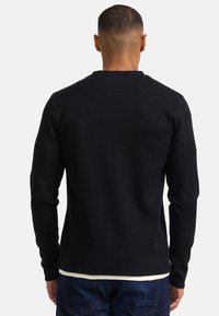 Camisa negra de manga larga con textura acanalada, diseño de cuello redondo con costuras sutiles, superpuesta sobre una camisa blanca, combinada con vaqueros azules.