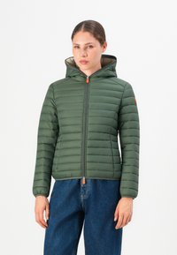 Vrouw in een groene gevoerde hooded jack met oranje ritsen en blauwe jeans, recht van voren tegen een witte achtergrond.