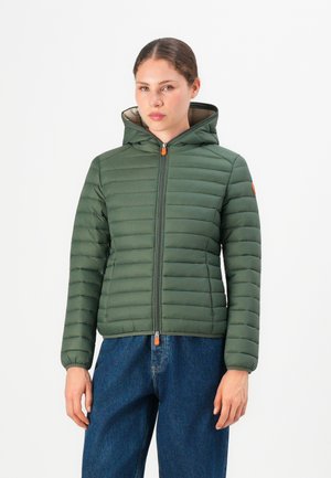 Vrouw in een groene gevoerde hooded jack met oranje ritsen en blauwe jeans, recht van voren tegen een witte achtergrond.