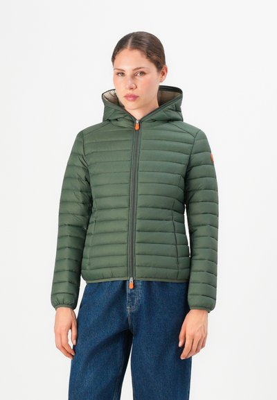 Femme portant une veste matelassée à capuche verte avec des tirettes orange et un jean bleu, de face contre un fond blanc.