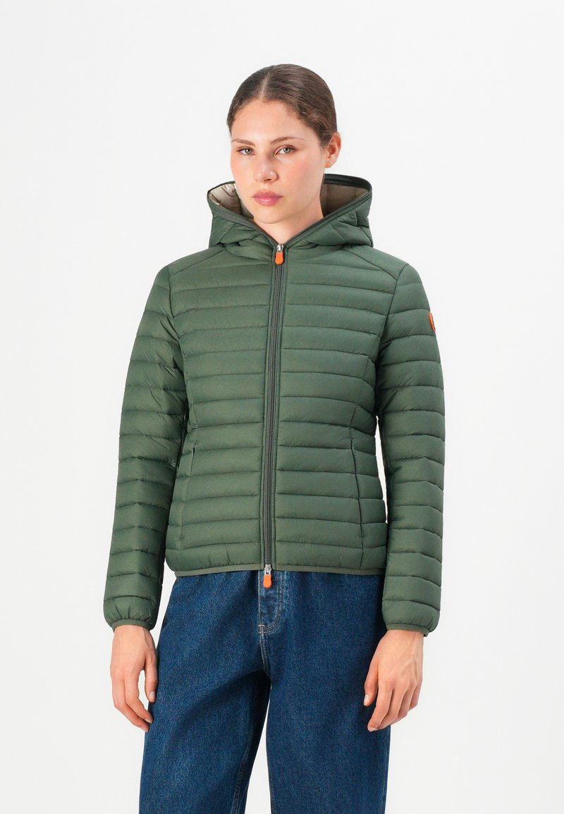 Vrouw in een groene gevoerde hooded jack met oranje ritsen en blauwe jeans, recht van voren tegen een witte achtergrond.