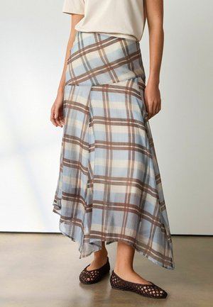 Frau trägt einen asymmetrischen Midi-Rock in blau-braidem Karomuster, kombiniert mit dunklen, gewebten Ballerinas auf einem glatten Boden.