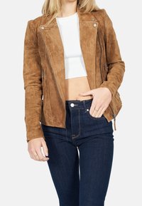 Donna che indossa una giacca con zip in suede marrone sopra un crop top bianco e jeans scuri a vita alta, con la mano che riposa nella tasca.