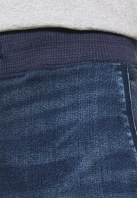 Närbild av midjebandet på blå denimjeans med ett mörkblått ribbat elastiskt band och en ljusgrå skjortkant ovanför.