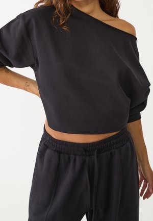Vrouw draagt zwarte off-shoulder cropped sweatshirt en zwarte sweatpants met elastische taille, hand op de heup en losse krullen zichtbaar.