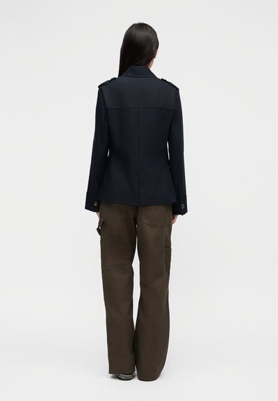 Γυναίκα με μακριά σκούρα μαλλιά, φοράει εφαρμοστή μαύρη ζακέτα και χαλαρό καφέ παντελόνι με τσέπες, στέκεται γυρισμένη προς τα πίσω σε απλό λευκό φόντο.