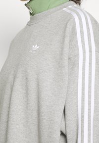 Grå sweatshirt med vita ränder på ärmarna, tillverkad av mjukt material. Har en liten vit logotyp på bröstet och en avslappnad passform.