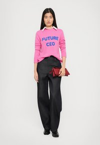 Rosa genser med "FUTURE CEO" i blått, hvit skjorte under, mørk brede bukser, holder en rød clutch, svarte sko.