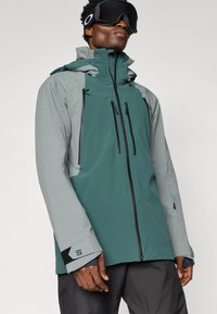 Férfi, aki kétféle zöld vízálló dzsekit és fekete sígödröt visel, egy egyszerű világos háttér előtt pózol.