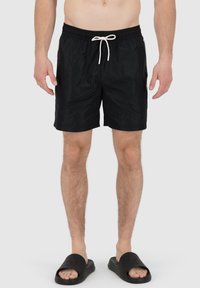 Pantaloni da bagno neri con cintura elastica e cordino bianco; materiale leggero; indossati con sandali neri.