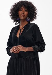 Czarna welurowa sukienka z długimi rękawami, z dekoltem na sznurek, teksturowymi mankietami oraz marszczonym dołem. Modelka nosi oversize'owe kolczyki.