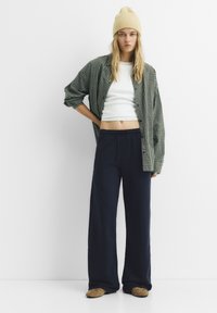 Karierte grüne Oversized-Hemdkombination, weißes Crop-Top, marineblaue weit geschnittene Hose, beigefarbene Mütze und gemusterte Slipper. Lässige Ensemble.