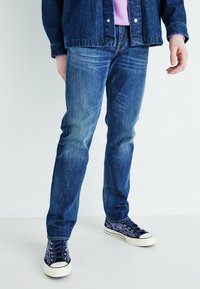 Keskmise sinise värvusega sirged denim püksid, kombineeritud tumesiniste tossudega, millel on leegimuster ja valge kummitald.
