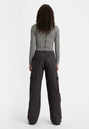 Personne aux cheveux bouclés portant un haut fleuri à manches longues et un pantalon cargo noir ample, debout, vue de dos sur fond blanc.