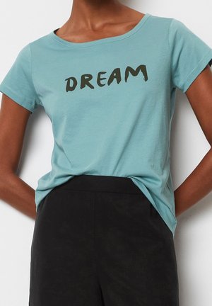 Frau trägt ein türkisfarbenes Kurzarmshirt mit schwarzem "DREAM"-Schriftzug und hochtaillierte schwarze Hose, Hände in die Hüften gestützt.