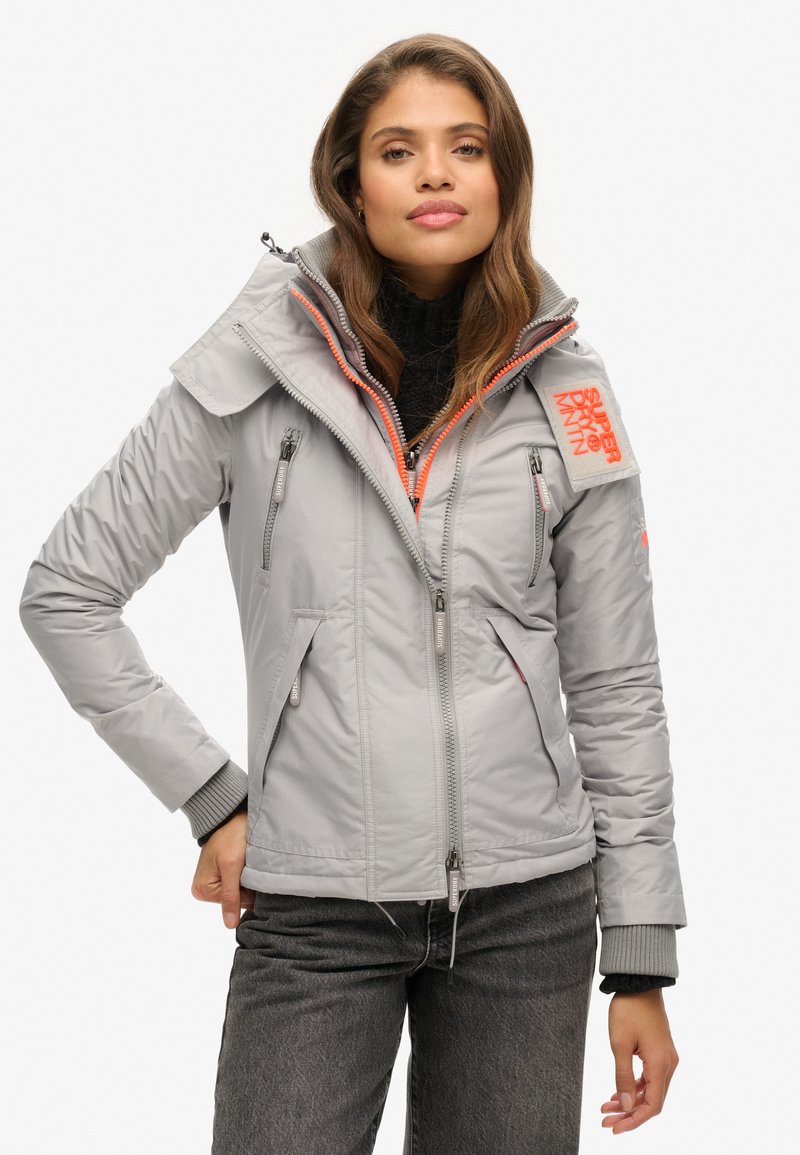 Veste isolée gris clair avec un col montant, accents de fermeture éclair orange et plusieurs poches. Présente une finition texturée et des poignets côtelés.