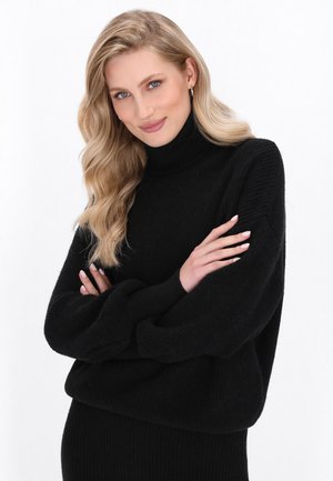 Czarny sweter z prążkowanym kołnierzem i luźnym krojem, z długimi rękawami. Miękka faktura i minimalistyczny design, noszony z rękami skrzyżowanymi na białym tle.