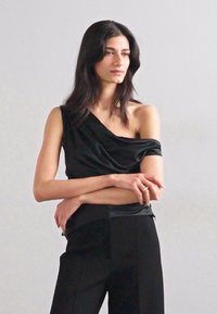 Fekete, egyvállas, redőzött dizájnnal készült felső, sima anyagból, testreszabott fekete nadrággal párosítva. Az öltözet minimalista stílusban készült.