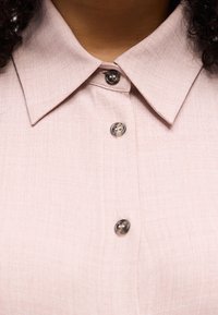 Κοντινό πλάνο ανοιχτόχρωμου ροζ πουκάμισου με γιακά και τρία σκούρα κουμπιά κλεισμένα, που δείχνει υφή υφάσματος και τμήμα του λαιμού και των μαλλιών.