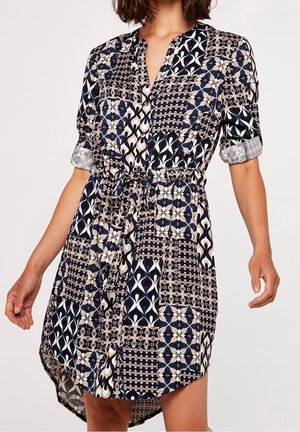 Femme portant une robe à motifs avec des manches retroussées, un col en V et une ceinture nouée, présentant des motifs géométriques bleu marine, beige et blanc.