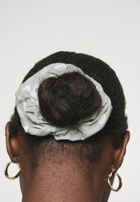 Scrunchie verde claro con lunares blancos asegurada alrededor de un moño rizado oscuro, usado en cabello corto y oscuro. Pendiente de aro dorado visible.