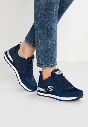 Ben i blå acid-wash jeans iført marineblå Skechers sneakers med hvide og sølv accenter, stående på en lysegrå gulv.