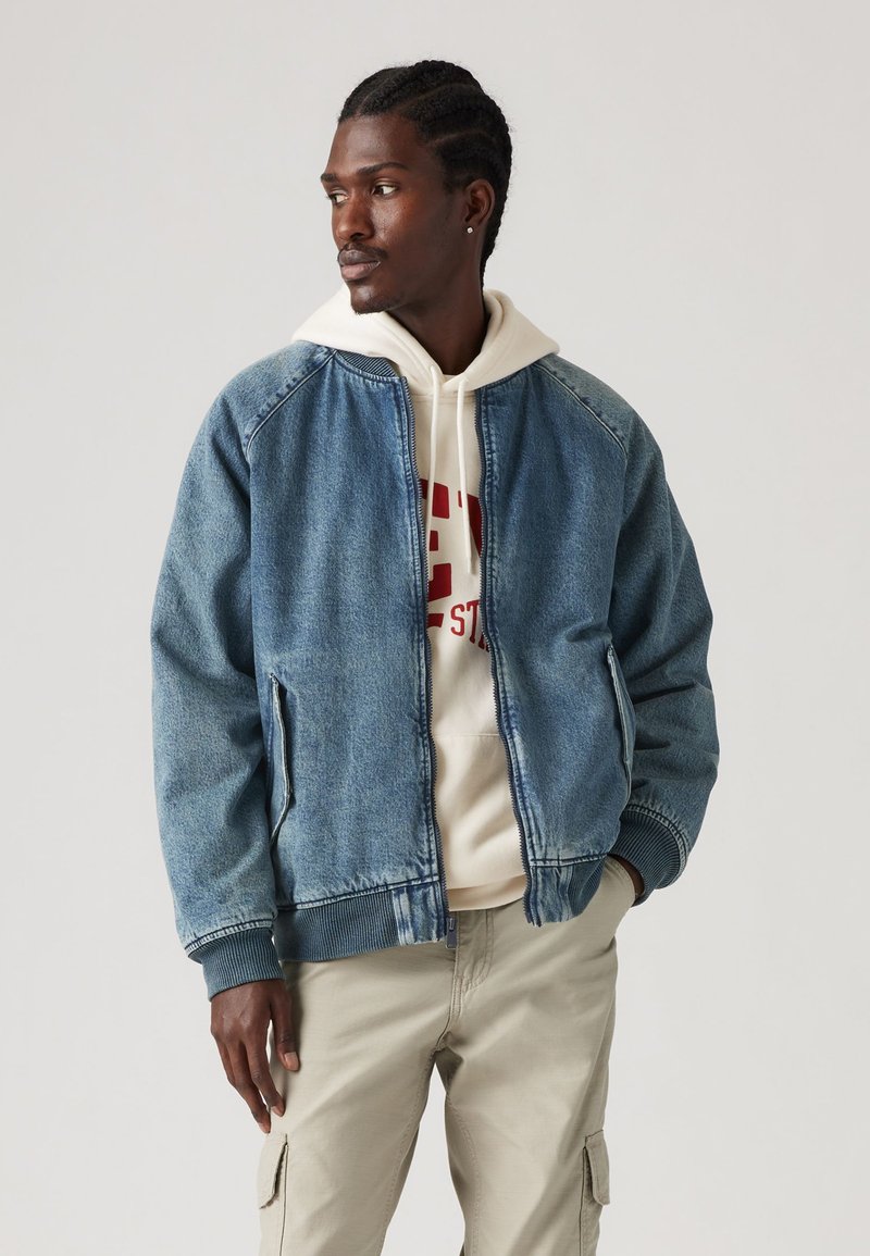 Chaqueta bomber de denim en azul claro, con puños y cinturilla acanalados. Lleva puesta una sudadera color crema con texto gráfico rojo. Pantalones color crema.