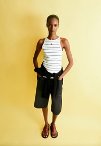 Gestreiftes weißes Tanktop mit einem roten Emblem, schwarze Shorts und schwarze Sandalen mit roten Akzenten, vor einem gelben Hintergrund.