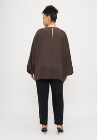 Brązowa bluzka z długim rękawem i tyłem w kształcie łezki, luźna, w połączeniu z czarnymi, dopasowanymi spodniami i czarnymi butami na obcasie.