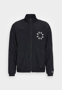 Svart lättviktsjacka med hög krage, dragkedjestängning, elastiska ärmslut och ett cirkulärt vitt grafiskt mönster på vänster bröst.