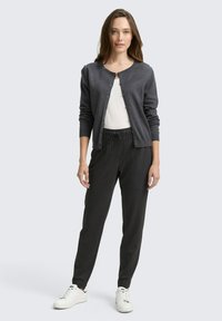 Grijze cardigan met ronde hals, knoopsluiting, gedragen over een witte top. Gecombineerd met zwart gestreepte broek en witte sneakers.