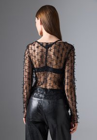 Top lungo a maniche sheer nere con ricami floreali, indossato sopra un canottiera nera. Abbinato a pantaloni in pelle neri a vita alta.