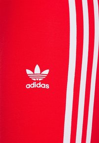 Röda sportleggings i stretchmaterial, med tre vita vertikala ränder längs sidan och en vit Adidas-logotyp längst ner.