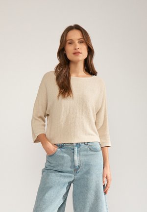 Vrouw met lang bruin haar die een beige textuurtrui met driekwartsmouwen en lichtblauwe, hooggetailleerde jeans draagt, staand met één hand in de zak.