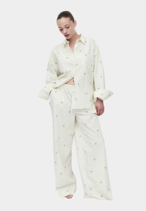 Kvinde iført løstsiddende, lyse farvet pyjamassæt med små blomsterprint, stående med den ene hånd i lommen og hovedet let nedadvendt.