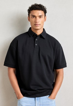 Black polo shirt made of soft fabric, featuring a classic collar, three-button placket, and short sleeves. Worn with light blue jeans.