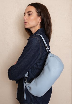 Lichtblauwe leren tas met een gebogen vorm, verstelbare schouderriem en zilverkleurige hardware, gedragen op de schouder over een donkere spijkerjack.