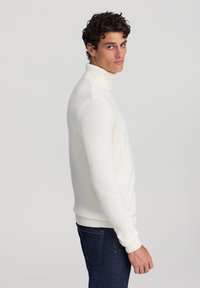 Maglione bianco a collo alto con orlo e polsini a costine, design a maglia testurizzata, abbinato a jeans blu scuro. Il modello è in profilo.
