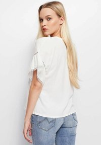 Camicia bianca con maniche bordate in pizzo e dettagli argentati, abbinata a jeans blu chiaro. Tessuto morbido con una vestibilità comoda.