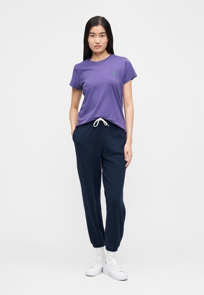 Lila rövid ujjú pamut póló zöld logóval, sötétkék jogger nadrággal és fehér tornacipővel párosítva. A nadrág rugalmas szegélyű.
