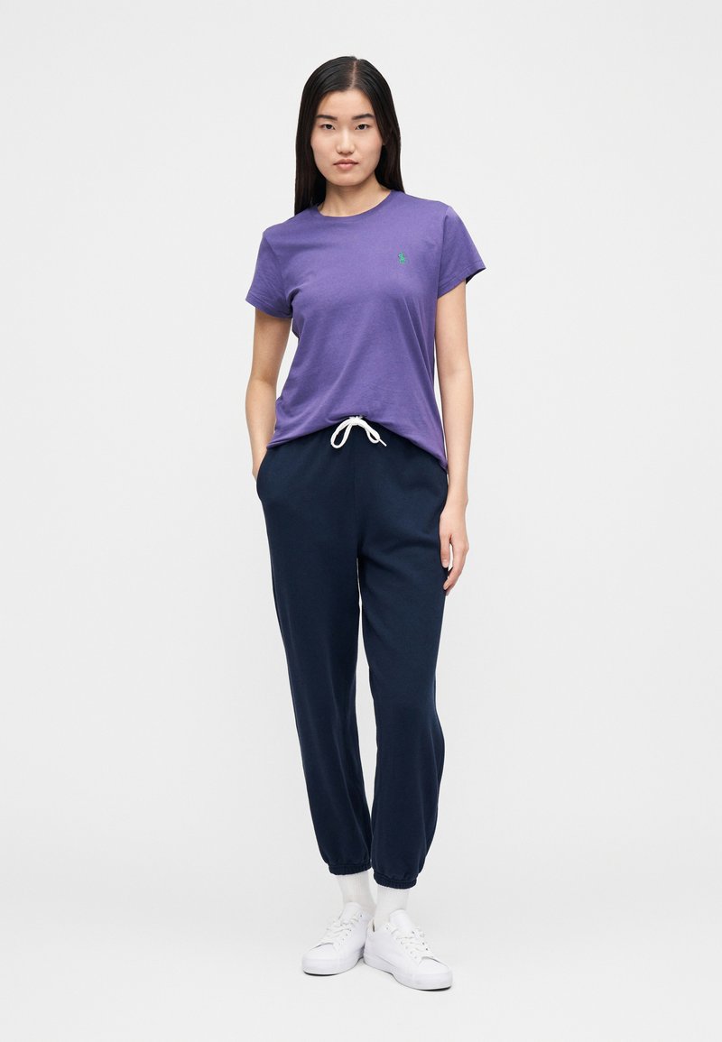 T-shirt en coton violet à manches courtes, avec un logo vert, assorti à un pantalon de jogging bleu marine et des baskets blanches. Le pantalon possède des poignets élastiques.