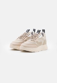 Beige tennarit, joissa on teksturoitu päällinen, pyöristetyt varpaat ja paksu kumipohja. Varustettu nauhakiinnityksellä ja mustilla logo-tunnuksilla kantapäässä.