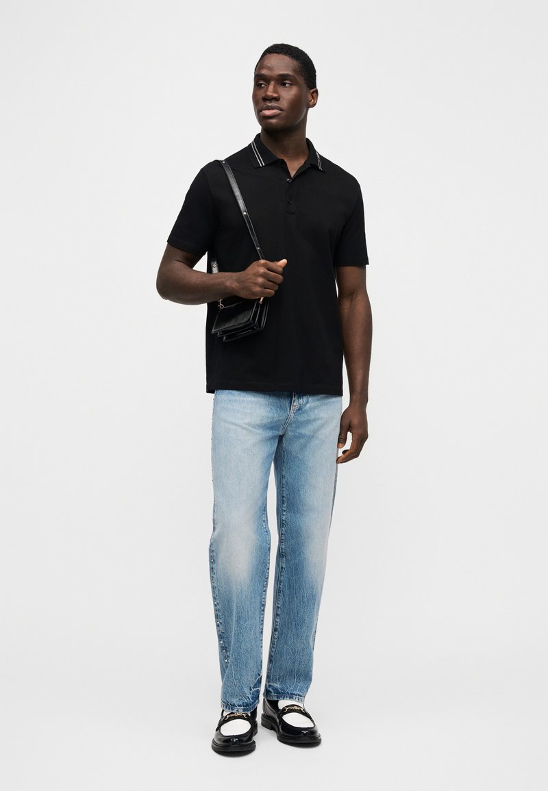 Homme debout portant un polo noir, un jean bleu clair avec des boutons latéraux, tenant un sac bandoulière noir, portant des mocassins noirs et blancs, sur fond blanc.