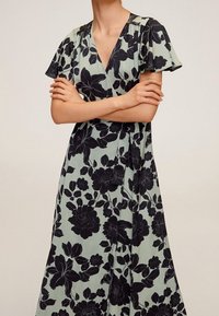 Robe portefeuille florale en tissu vert clair ornée de motifs floraux noirs, avec des manches courtes et flottantes, et un décolleté en v avec une taille cintrée.