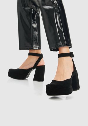 Jambes portant un pantalon court en cuir verni noir et des talons blocs plateformes en suède noir avec des brides à la cheville sur fond blanc.