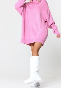 Nadměrný růžový pletený svetr s vysokým límcem, se žebrovanými manžetami a lemem, doplněný vysokými bílými gumovými botami.